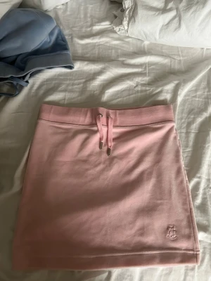 Ljusrosa kortkjol med snörning - Säljer en ljusrosa kortkjol från Juicy Couture med snörning i midjan och broderad logga längst ner. Kjolen är gjord i mjukt syntetmaterial och har en enkel, sportig design som är perfekt för sommarens alla äventyr.