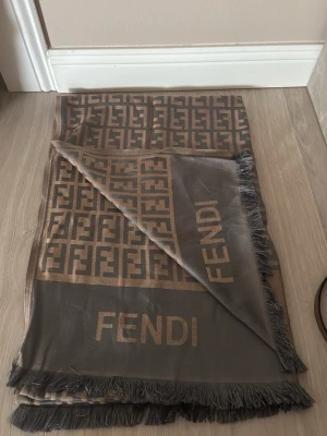 Brun och beige sjal från Fendi - Snygg sjal från Fendi i brunt och beige med klassiskt FF-mönster och logotyp. Sjalen har fransade kanter och är tillverkad i ett mjukt material som känns lyxigt. Perfekt accessoar för att lyfta din outfit.