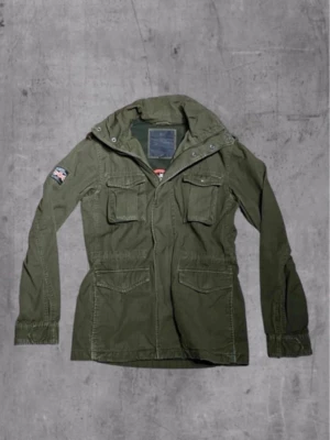 Olivgrön parkas från Superdry - Säljer en olivgrön parkas från Superdry med fyra stora fickor framtill, huva och patch med brittisk flagga på ärmen. Jackan har dragkedja och tryckknappar samt justerbar midja. Tillverkad i slitstarkt bomullstyg, perfekt för en cool streetstil.