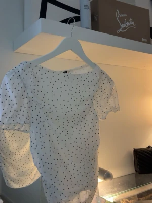 Vit prickig blus med puffärm - Supersöt vit blus med svarta prickar och puffiga, transparenta ärmar. Blusen har en feminin passform och är gjord i ett lätt, luftigt material som känns perfekt för varmare dagar. Perfekt att styla med jeans eller kjol för en trendig look.