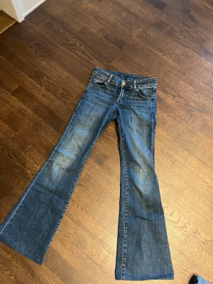 Blå bootcut jeans med slitningar - Säljer ett par blå bootcut jeans med snygga slitningar och klassiska fem fickor. Jeansen har låg midja och är tillverkade i denim med kontrastsömmar. Perfekta för en avslappnad och trendig look.