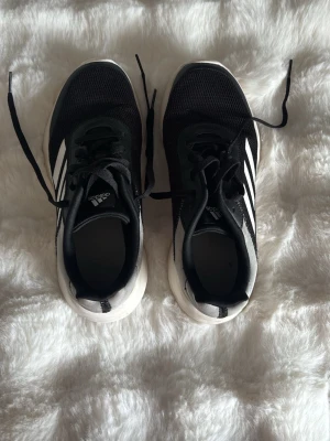 Svarta Adidas sneakers med vita ränder - Snygga svarta Adidas sneakers med klassiska vita ränder på sidorna och vit sula. Ovandelen är i mesh-tyg för bra andningsförmåga och skorna har svarta skosnören. Perfekta för en sportig och avslappnad stil.