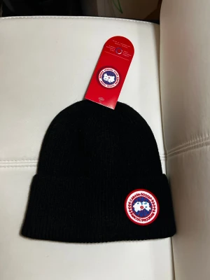 Svart mössa från Canada Goose - Säljer en svart ribbstickad mössa från Canada Goose med klassisk rund logotyp framtill. Mössan har uppvikt kant och är tillverkad i mjukt material som håller värmen. Perfekt accessoar för kalla dagar och snygg till alla outfits.