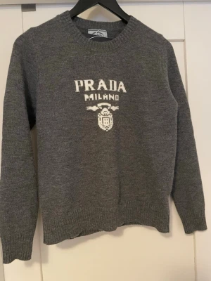 Grå stickad tröja från Prada - Grå stickad tröja från Prada med vit logotyp och texten 'PRADA MILANO' framtill. Tröjan har rund halsringning, långa ärmar och ribbstickade muddar vid ärmslut och nederkant. Perfekt för dig som gillar exklusiva märken och stilren design.