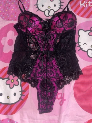 Svart och rosa spetsbody med snörning - Säljer en snygg body i svart spets med rosa underton. Bodyn har tunna axelband, vadderade kupor och dekorativ snörning framtill. Spetsdetaljer och öppen rygg ger en extra touch. Perfekt för dig som vill ha något unikt och vågat.