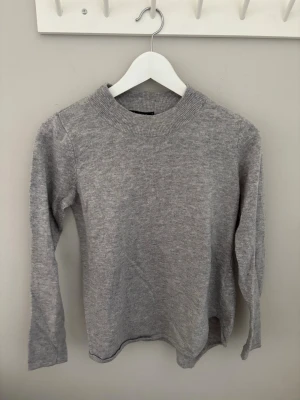 Ljusgrå stickad tröja - En enkel och stilren ljusgrå stickad tröja med rund hals och långa ärmar. Tröjan har ribbstickade muddar vid halsen och är tillverkad i ett mjukt material som passar perfekt till lager på lager. Perfekt basplagg för kyligare dagar.