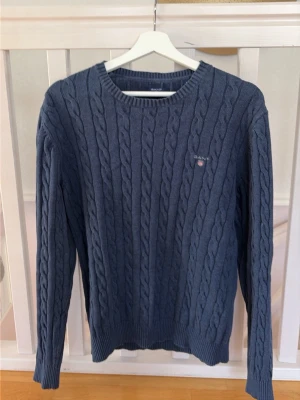 Blå kabelstickad tröja från Gant - Kabelstickad blå tröja från Gant med rund halsringning och diskret logga broderad på bröstet. Tröjan har långa ärmar och ribbade muddar vid ärmslut och nederkant. Perfekt för dig som gillar klassisk och stilren design.