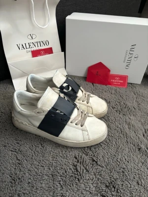 Valentino Garavani vita sneakers - Snygga vita sneakers från Valentino Garavani med en bred svart läderdetalj över sidan. Skorna har klassisk låg profil, vita snören och diskret logga på tungan. Tillverkade i läder för en lyxig känsla och stilren look. Pris kan diskuteras! 