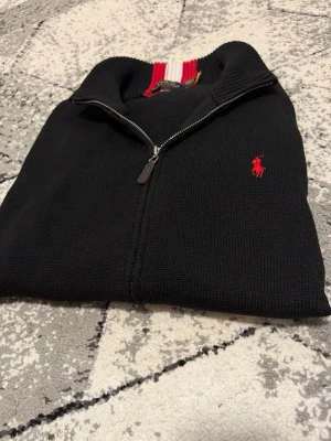 Svart fullzip från Polo Ralph Lauren - Svart stickad tröja med  dragkedja från Polo Ralph Lauren. Klassisk röd logga broderad på bröstet och ribbad krage med röd och vit detalj på insidan. Perfekt för en stilren och avslappnad look.