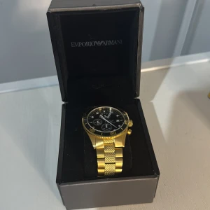 Guldfärgad klocka från Emporio Armani - Stilren guldfärgad klocka från Emporio Armani med svart urtavla och kronograf. Klockan har en robust länkarmband i metall och tydliga markeringar på boetten. Perfekt accessoar för dig som vill sticka ut med en lyxig look. DENNA KLOCKAN ÄR ÄKTA‼️