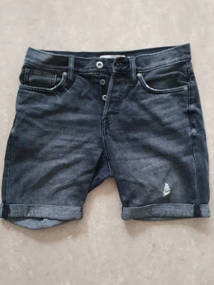 Jeansshorts - Snygga svarta jeansshorts med uppvikta benslut och knappgylf. Klassisk femficksmodell med en liten sliten detalj framtill. Perfekta för en avslappnad stil under varma dagar. 29 tum.