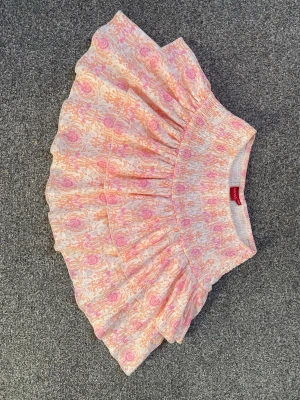 Mönstrad volangkjol från RYVLS - Superfin kort volangkjol från RYVLS med rosa och orange mönster på vit botten. Kjolen har bred smock i midjan och flera lager volanger som ger en riktigt söt och lekfull look. Perfekt för dig som gillar färg och mönster!