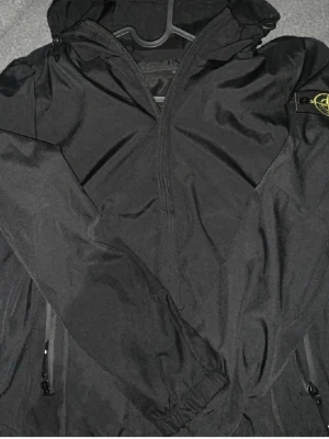 Svart vindjacka från Stone Island - Snygg svart vindjacka från Stone Island med huva och dragkedja framtill. Jackan har två sidofickor med dragkedja och den klassiska Stone Island-loggan på ärmen. Tillverkad i ett lätt och slitstarkt material med slät insida. Helt Ny!