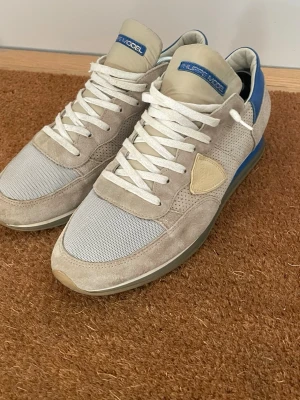 Philippe Model beige sneakers - Snygga sneakers från Philippe Model i beige mocka och mesh med vita skosnören och blå detaljer vid hälen. Skorna har en låg silhuett och en gummisula med fiskbensmönster. Perfekta för en avslappnad och trendig stil.