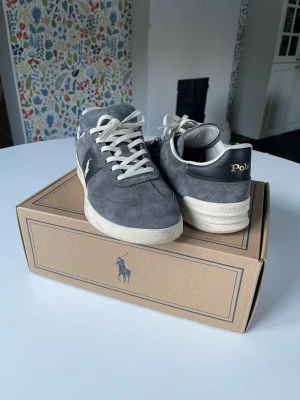 Blå/Grå Polo Ralph Lauren Heritage Aera Suede Sneakers - Snygga sneakers i grå mocka från Polo Ralph Lauren med vita snören och vit sula. Klassisk låg modell med Polo-logga i guld på hälen och broderad häst på sidan. Dem var för små för mig och bara använda typ 5 ggr.