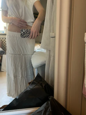 Vit långkjol med volangdetaljer - Säljer en vit långkjol med volangdetaljer och lätt skrynklig look. Kjolen har en avslappnad passform och är perfekt för varmare dagar. Tillverkad i ett tunt och mjukt material som känns luftigt och bekvämt. Se sista bilden för defekter.