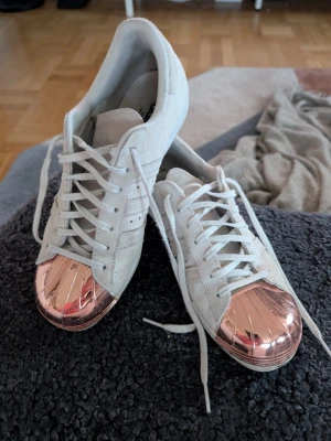 Vita Adidas sneakers med kopparfärgad tå - Säljer ett par vita Adidas sneakers med klassiska ränder på sidan och en unik kopparfärgad tå i metall. Skorna har vita snören och sula i gummi. Perfekta för dig som vill sticka ut med en cool detalj på fötterna.