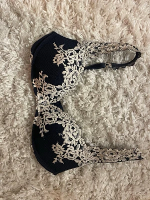 Mörkblå spets-bh med blommig brodyr - Snygg mörkblå bh med vadderade kupor och vacker beige spetsbrodyr i blommigt mönster. Axelbanden är dekorerade med samma spets och bh:n har hakknäppning bak. Perfekt för dig som gillar romantiska detaljer.