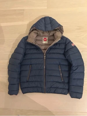 Colmar dunjacka - Snygg blå dunjacka från Colmar Originals med beige insida och vadderad design. Jackan har två dragkedjeförsedda fickor, huva och Colmar-logga på ärmen. Perfekt för kalla vinterdagar och riktigt skön att bära. I storlek M