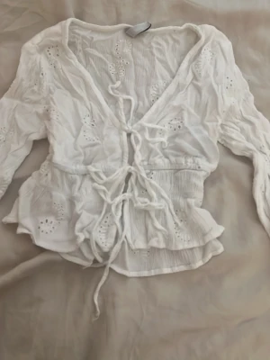 Vit broderad blus med knytning - Söt vit blus med broderade detaljer och knytning framtill. Blusen har v-ringning, volangkant nedtill och långa ärmar med lätt puff. Materialet är tunt och luftigt, perfekt för sommardagar. Ger en romantisk och bohemisk vibe.