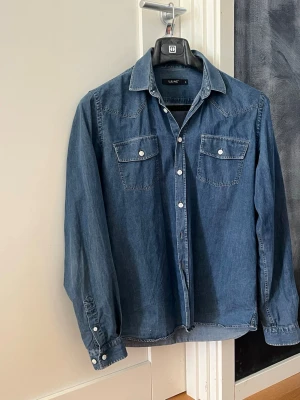 Blå jeansskjorta från Ljung - Säljer en klassisk blå jeansskjorta från Ljung med två bröstfickor och vita knappar. Skjortan har lång ärm och en avslappnad passform, perfekt till jeans eller chinos. Materialet är mjukt och slitstarkt denim i bomull.