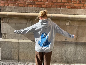 Grå hoodie med blått tryck -  en grå hoodie med stor huva och ett blått grafiskt tryck på ryggen där det står ' Keep Fashion Weird'. Hoodien har en avslappnad passform. Perfekt för dig som gillar trender och vill sticka ut lite extra.