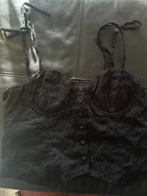 Svart broderad korsettopp  - Snygg svart korsettopp från NA-KD i storlek 40 med broderade detaljer och knappar framtill. Toppen har tunna axelband som knyts själv. 🪩🪩