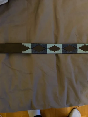 Brunt skärp med mönster i skinn - Snyggt brunt skärp i skinn med geometriskt mönster i beige och mörkgrått. Bältet har bred design och detaljer som ger en unik look. Perfekt accessoar för att lyfta din outfit.