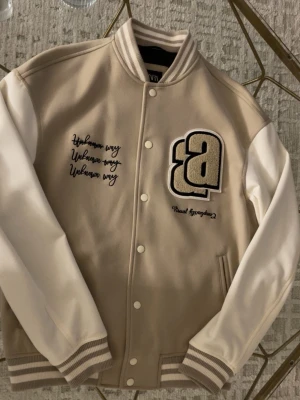 Beige varsityjacka - Snygg beige och vit jacka från ZARA med broderade detaljer och texten 'Unknown way' samt stor bokstavslogga på bröstet. Jackan har ribbade muddar, tryckknappar och kontrasterande ärmar. Helt ny skick fläckfri använd 1 gång och sälj pga det ej passar min stil riktigt.
