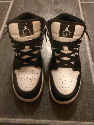 Nike Air Jordan 1 Mid svart/vit - Snygga Nike Air Jordan 1 Mid sneakers i storlek 38 de är svart och vitt. Klassisk design med svart swoosh, perforerad tåbox och högt skaft. Skorna har svarta skosnören och är tillverkade i läder och syntetmaterial. Perfekta för dig som gillar streetwear och ikoniska sneakers. (Sko lådan följer inte med)