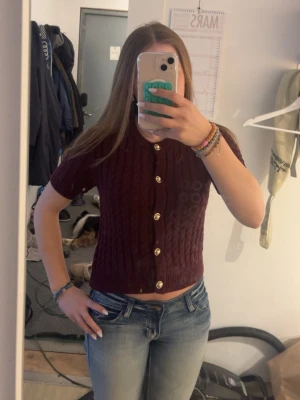 Vinröd kabelstickad kortärmad kofta - Kortärmad vinröd kofta med kabelstickat mönster och dekorativa guldknappar framtill. Modellen är croppad och har rund hals. Perfekt att styla med jeans eller kjol för en trendig look. Aldrig använt lappen fortfarande kvar