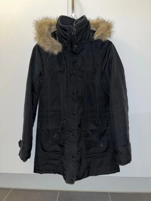 Svart parkas med fuskpäls - Snygg svart parkas med avtagbar huva och fuskpäls runt kanten. Jackan har dragkedja och knappar framtill, fyra fickor med lock och muddar vid ärmsluten. Varm och skön, Perfekt för kalla dagar. Klassisk modell som funkar till det mesta.