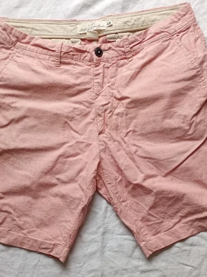 Ljusrosa shorts från H&M - Säljer ett par ljusrosa shorts från H&M med klassisk passform och knappgylf. Shortsen har bälteshällor, sidofickor och är tillverkade i mjuk bomull. Perfekta för varma dagar och enkel att matcha med olika toppar.