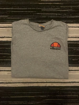 Grå t-shirt från Ellesse - Säljer en stilren grå t-shirt från Ellesse med klassisk logga i orange och rött på bröstet. T-shirten har rund hals och korta ärmar, perfekt för en avslappnad look. Tillverkad i mjuk bomull för skön komfort.