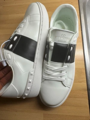 Valentino Garavani vita sneakers - Snygga vita sneakers från Valentino Garavani med bred svart läderdetalj och nitar på sidan. Skorna har vita snören, platt sula med mönster och logga på plösen. Perfekta för dig som gillar stilrena och exklusiva sneakers.