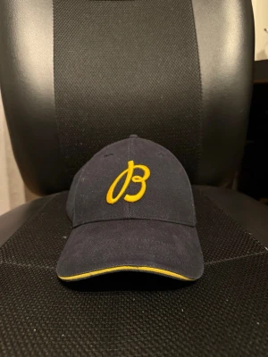 Mörkblå keps från Breitling - Snygg mörkblå keps från Breitling med gul broderad logga framtill och justerbart läderband bak. Kepsen har gul detalj längs skärmen och är tillverkad i ett tåligt tygmaterial. Perfekt accessoar för dig som gillar stilrena detaljer och exklusiva märken.