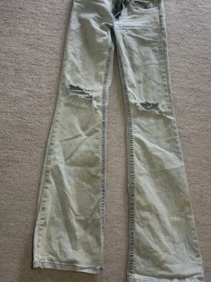 Ljusblå bootcut jeans med håll - Säljer ett par ljusblå bootcut jeans med slitna detaljer över knäna och en cool röd broderad detalj på bakfickan. Jeansen har klassisk femficksdesign och är tillverkade i denim. Perfekta för en avslappnad och trendig look.