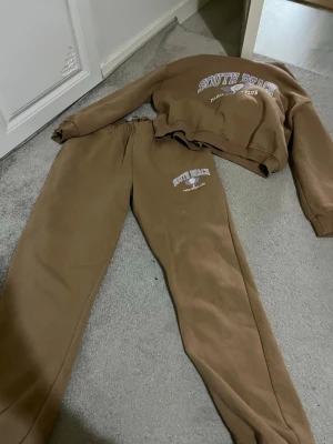 Bruna mjukisbyxor South Beach - Bruna mjukisbyxor från South Beach med tryckt logga och text på ena benet. Byxorna har resår i midjan och är gjorda i mjukt bomullsmaterial. Perfekta för en avslappnad och chill stil.