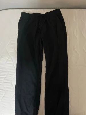 Svarta mjukisbyxor med dragsko - Säljer ett par svarta mjukisbyxor med resår i midjan och dragsko för skön passform. Byxorna har raka ben och fickor både fram och bak. Perfekta för chill dagar eller när du vill vara bekväm. Materialet känns mjukt och följsamt.