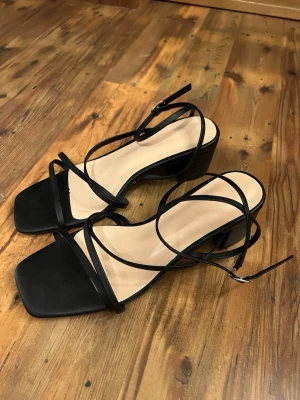Svarta sandaler med blockklack och remmar - Säljer ett par stilrena svarta sandaler med tunna remmar och fyrkantig tå. Skorna har en bred blockklack och justerbart spänne vid ankeln. Materialet ser ut att vara konstläder och designen är minimalistisk och trendig. klacken är 5cm, aldrig använda. 