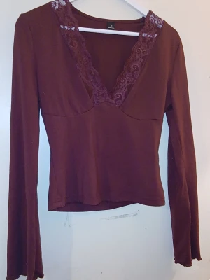 Brun långärmad topp med spets - Snygg brun långärmad topp med djup v-ringning och dekorativ spets längs urringningen. Figurnära passform och mjukt material som känns skönt mot huden. Perfekt för dig som vill ha en enkel men ändå detaljrik topp.
