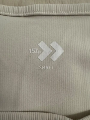 Vit långärmad topp från 157 - Säljer en stilren vit långärmad topp från 157 med ribbad struktur och rundad halsringning. Toppen har en figurnära passform och är perfekt att matcha med jeans eller kjol. Diskret logga nertill på sidan.