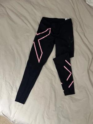 Svarta 2XU leggings med rosa detaljer - Säljer ett par svarta leggings från 2XU med snygga rosa mönster längs benen. De har hög midja och är tillverkade i ett stretchigt funktionsmaterial som passar perfekt till träning. Loggan syns diskret på låret. Supersnygga och bekväma tights!