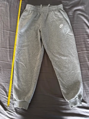 Gråa Nike mjukisbyxor - Gråa mjukisbyxor från Nike med resår i midjan och dragsko för skön passform. Byxorna har muddar vid bensluten och sidofickor. Tillverkade i mjukt bomullsmaterial och har en vit Nike-logga broderad på vänster ben. Perfekta för träning eller chill. Storleken 158/170(för dig som är ca 160 lång)