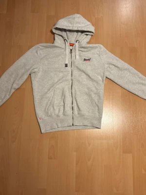 Ljusgrå hoodie från Superdry - Ljusgrå zip-hoodie från Superdry med broderad logga på bröstet. Hoodien har dragsko i huvan, två fickor fram och ribbade muddar vid ärmslut och nederkant. Perfekt för en avslappnad och stilren look. Storlek L i barn men jag använder S och passar mig perfekt 