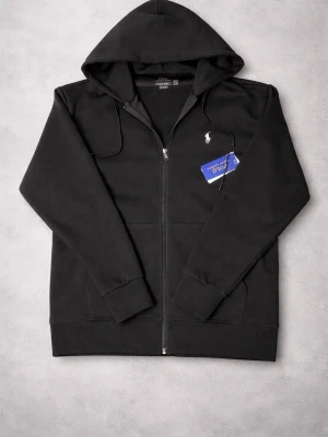 Svart hoodie från Polo Ralph Lauren - Snygg svart hoodie med dragkedja från Polo Ralph Lauren. Klassisk design med vit broderad logga på bröstet, huva med dragsko och två fickor framtill. Tillverkad i mjukt bomullsmaterial som är perfekt för en chill och stilren look.