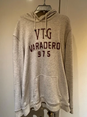  hoodie från Jack & Jones - Säljer en beige hoodie från Jack & Jones Vintage Cloth med texten 'VTG VARADERO 975' i vinrött framtill. Hoodien har huva med dragsko, magficka och är gjord av återvunnen bomull. Perfekt för en avslappnad och trendig stil.
