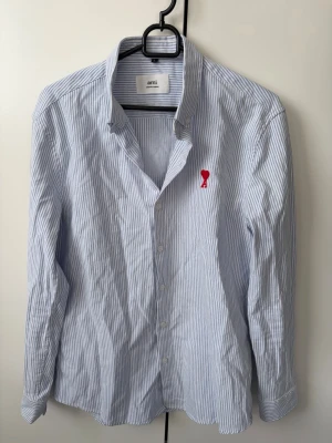 Blårandig skjorta från Ami Paris - Säljer en klassisk blårandig skjorta från Ami Paris med röd broderad logga på bröstet. Skjortan har lång ärm, knappar framtill och är gjord i mjuk bomull. Perfekt för en clean och stilren look. Storlek L men sitter som en M kan passa S.