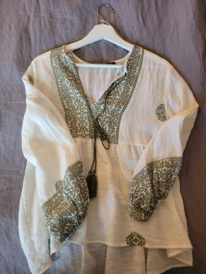 Vit boho-blus med grönt mönster Zara - Bohemisk vit blus från Zara med grönt broderat mönster längs v-ringningen och ärmsluten. Blusen har långa, luftiga ärmar och dekorativ knytning med tofs framtill. Tillverkad i ett tunt, vävt material som ger en avslappnad och trendig look.