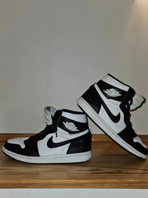 Air Jordan 1 High svart/vit - Säljer ett par klassiska Air Jordan 1 High sneakers i svart och vitt läder. Skorna har den ikoniska Nike Swoosh-loggan på sidan och Air Jordan Wings-märket vid ankeln. Hög modell med snörning och vadderad plös för extra komfort. Har hål vid hälarna